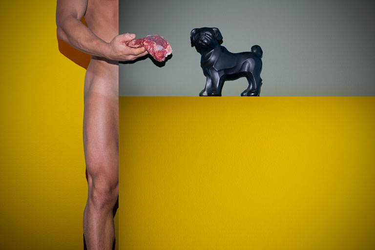Félix Ossa - Doméstico - Imagen de el cuerpo de un hombre desnudo detrás de una pared. El hombre le ofrece carne a un animal de adorno